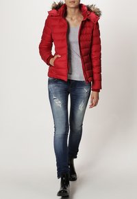 Rød pufferjakke med hætte og faux fur-kant, lynlåslukning og sidelommer, båret over en grå t-shirt og slidte denimjeans.