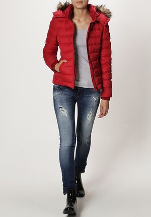 Rote Daunenjacke mit Kapuze und einem Besatz aus Kunstpelz, Reißverschluss und Seitentaschen, getragen über einem grauen Shirt und verwaschenen Jeans.