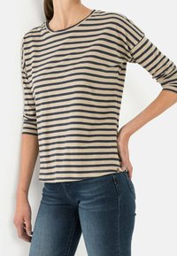 Woman wearing beige and black horizontal striped long-sleeve shirt and dark blue jeans, hand resting on hip.