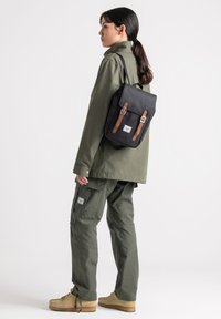 Black backpack with brown leather straps, rectangular shape, smooth texture, and a front pocket. Worn with olive green clothing.