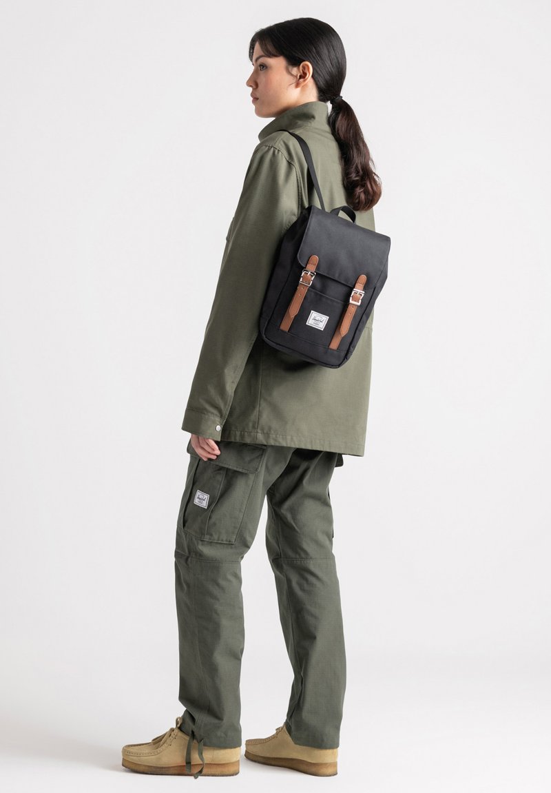 Black backpack with brown leather straps, rectangular shape, smooth texture, and a front pocket. Worn with olive green clothing.