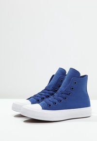 High-top sneakers in blue canvas with white rubber toe cap and sole. Features include six eyelets, flat laces, and textured surface.