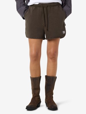 Person iført brune shorts med snøre og mørkebrune læderstøvler til midt på læggen, hænderne i lommene på shortsene, hvid baggrund.