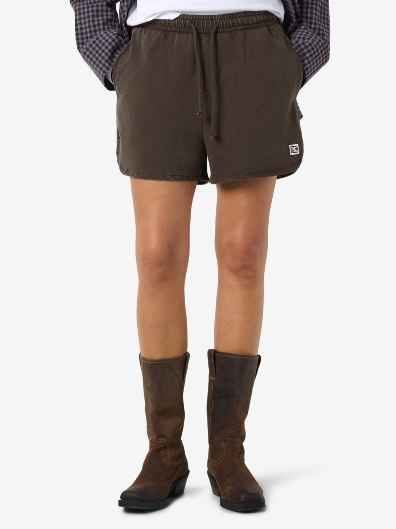 Άτομο που φοράει καφέ σορτς με κορδόνι και καφέ δερμάτινες μπότες μέσου μήκους, με τα χέρια στις τσέπες του σορτς, σε λευκό φόντο.