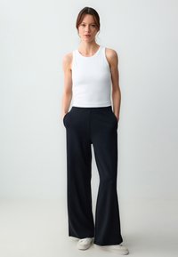 Witte mouwloze top gecombineerd met zwarte wijde broek. De broek heeft zijzakken en een gladde textuur. Model staat op een neutrale achtergrond.