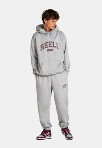Grauer Hoodie und Jogginghose mit maroonfarbener „REELL“-Schrift; weicher Stoff, lockere Passform, Fronttasche am Hoodie, elastische Bündchen an der Hose.