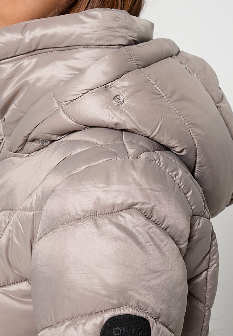 Primo piano di un giubbotto invernale trapuntato beige con cappuccio e una patch nera sulla manica indossato da una persona.