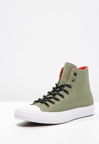 Sneaker alta realizzata in tessuto verde testurizzato con lacci neri, punta in gomma bianca e foderatura interna rossa.