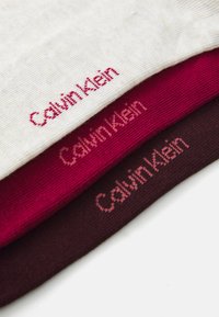 Three pairs of socks in white, red, and dark brown, featuring "Calvin Klein" logo in a contrasting color on each pair. Cotton blend texture.