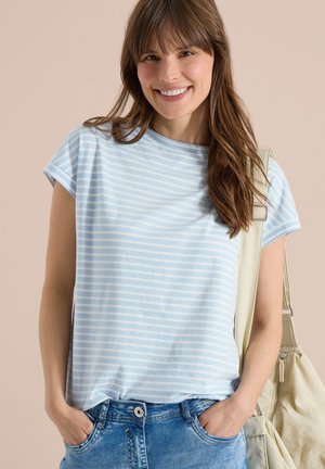 Ung kvinde smiler, iført en lyseblå og hvid stribet kortærmet skjorte, blå jeans, med hænderne i lommerne og en beige taske over skulderen.