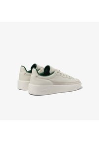 Pár nízkých tenisek v off-white barvě s zeleným vnitřním lemováním, velurovým bočním pruhem, šněrováním a s embosovaným nápisem "Lacoste" na podrážce.
