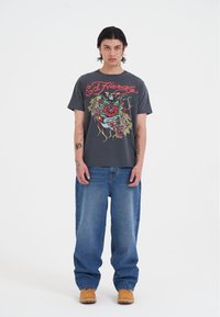 Camiseta de algodón gris con un gráfico colorido que presenta una rosa, un águila y texto. Llevada con jeans holgados azules y zapatos de lona marrones.