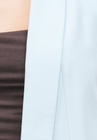 Svetlomodrý blejzer s hladkou tkaninou, štruktúrovaným dizajnom a otvoreným predným dielom. Nosený cez tmavohnedý top s ryhovanou textúrou.