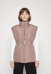 Colete acolchoado em rosa suave com gola alta, fecho de zip e cintura cingida; possui grandes aberturas para os braços e textura matizada discreta.