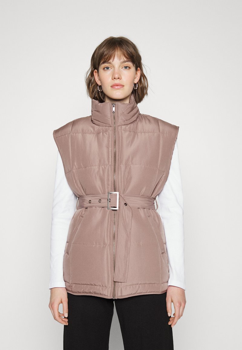 Colete acolchoado em rosa suave com gola alta, fecho de zip e cintura cingida; possui grandes aberturas para os braços e textura matizada discreta.