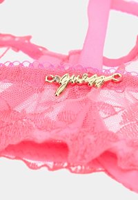 Pink lace bralette with ruffled edges, featuring a gold "guess" logo charm with a small rhinestone accent. Smooth straps in a matching color.