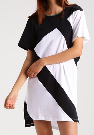 Femme portant une robe noire et blanche à manches courtes avec de larges blocs de couleur géométriques diagonaux sur un fond uni.