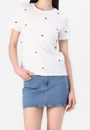 Vrouw draagt een wit T-shirt met ronde hals en kleine rode hartjes, gecombineerd met een blauwe denim minirok, hand in de zak.