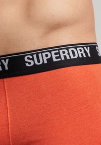 Boxer shorts de algodón naranja con una cinturilla elástica negra que presenta el nombre de la marca "SUPERDRY" en blanco. Vista en primer plano de la textura del tejido.
