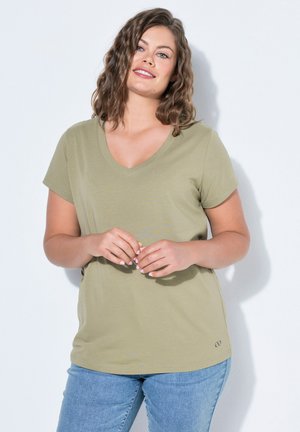 Frau mit welligem braunem Haar, die ein hellgrünes V-Ausschnitt-T-Shirt und blaue Jeans trägt, vor weißem Hintergrund stehend, lächelnd.