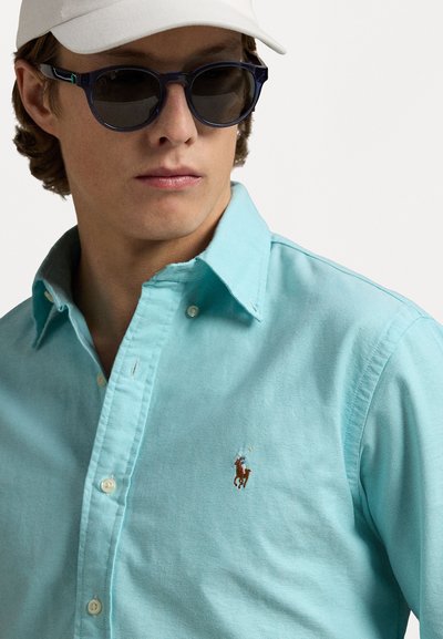 Gaiši zila krekls ar apkakli un pogām, ar izšūtu logo kreisajā krūšu daļā. Valkāts ar tumšām saules brillēm un baltu cepuri.