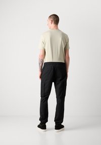 Zwarte cargobroek met twee zijzakken, elastische tailleband en boorden. Gecombineerd met een lichtbeige korte mouw t-shirt, getoond vanuit de achterkant.