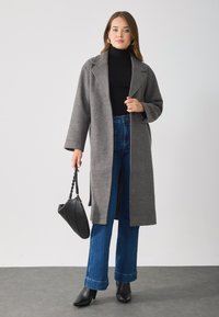 Grå ullkåpe med revers, lange ermer og rett snitt, parret med blå vidde jeans og svarte ankelstøvler. Holder en svart veske.