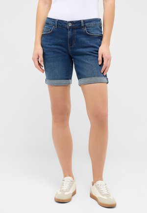 Frau trägt hochgekrempelte dunkelblaue Jeansshorts, weißes Oberteil und weiße Turnschuhe vor hellem, einfarbigem Hintergrund.