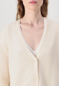 Beige cardigan med v-udskæring, der har en blød tekstur, tre knapper foran og subtil tonet detaljering langs udskæringen.
