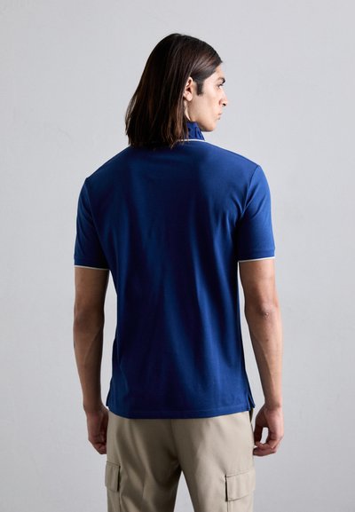 Bărbat cu păr brunet care atinge umerii, îmbrăcat cu un tricou polo albastru cu mâneci scurte și pantaloni cargo bej, stând cu fața spre un perete gri.