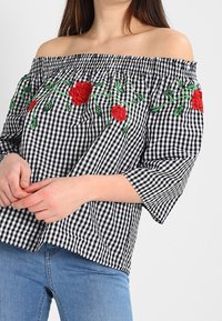 Blusa a scacchi in bianco e nero, con spalle scoperte, decorata con ricami floreali rossi sul petto e maniche ampie e svasate. Materiale in cotone.