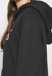 Sudadera negra con cordón, que presenta un logo en blanco y rojo. La tela es suave y lisa, con un corte holgado y un bolsillo tipo canguro.