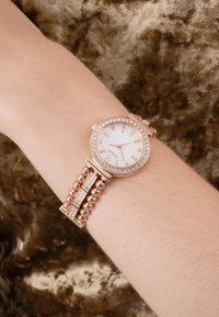 Różowa złota bransoletka z okrągłą tarczą, białą cyferblatą, elementami kryształowymi oraz bransoletą w formie koralików z prostokątnymi ogniwami.