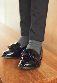 Sorte laksko med bånddetalje, båret med grå sokker, hvilende på en træoverflade. Skoene har rundet snude og lav hæl.