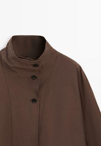 Brązowy płaszcz z wysokim kołnierzem, z trzema czarnymi guzikami ułożonymi pionowo w pobliżu kołnierza, prezentowany na białym tle.