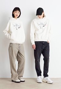 Sweatshirts blancs avec des impressions graphiques, associés à des jeans oversized beiges et noirs. Des baskets complètent les tenues. Design simple et décontracté.