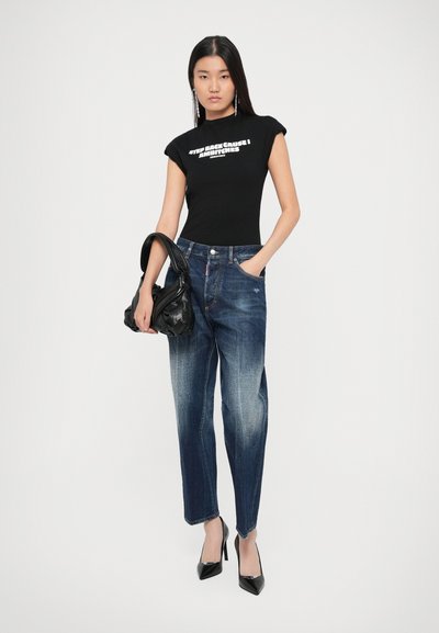Camiseta negra ajustada con texto blanco, jeans de mezclilla azul con detalles desgastados y zapatos de tacón negros. Bolso negro en la mano.