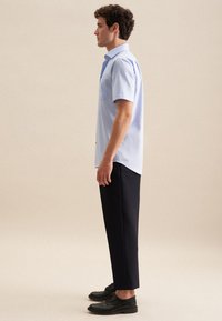 Camicia azzurra a maniche corte con una singola tasca sul petto, abbinata a pantaloni neri a gamba stretta e scarpe nere con lacci, posato di profilo.