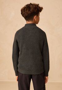 Maglione verde scuro lavorato a maglia con collo alto, trama a costine e polsini aderenti, visto da dietro su sfondo neutro.