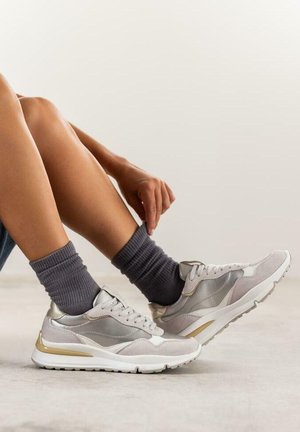 Zapatillas con parte superior de malla y gamuza en gris claro, acentos en dorado y suelas blancas. Combinadas con calcetines de canalé grises.