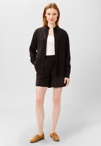 Černá košile s dlouhými rukávy a kapsou na hrudi, v kombinaci s černými šortkami s elastickým pasem a hnědými semišovými mokasíny.