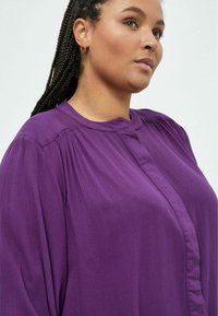Blusa viola con un colletto alto, dettagli plissettati e una chiusura a bottoni. Realizzata in un tessuto fluido, presenta una vestibilità comoda e una consistenza morbida.