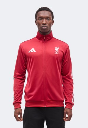 Άνδρας που φοράει κόκκινο ζακέτα Liverpool FC με λευκό λογότυπο Adidas και ρίγες στα μανίκια, στέκεται μπροστά σε ανοιχτό γκρι φόντο.