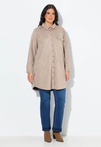 Beige Wildlederhemdkleid mit Knopfleiste, spitzem Kragen und einer Brusttasche. Kombiniert mit blauen Denim-Jeans und braunen Knöchelschuhen.