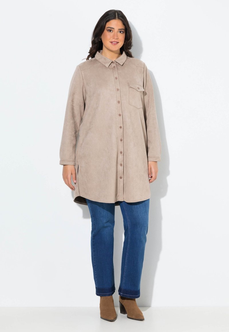 Beige Wildlederhemdkleid mit Knopfleiste, spitzem Kragen und einer Brusttasche. Kombiniert mit blauen Denim-Jeans und braunen Knöchelschuhen.