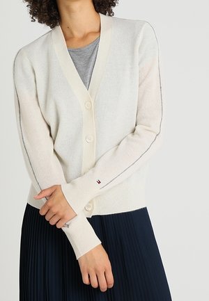 Cardigan en tricot crème clair avec un col en V, cinq boutons, coutures grises contrastées et poignets côtelés. Assortie avec une chemise grise et une jupe bleu marine.