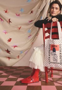 Červené lodičky z lesklé lakované kůže se špičatou špičkou a přezkami, doplněné červenými ponožkami, bílou plisovanou sukní a vzorovanou taškou přes rameno.