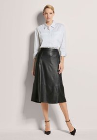 Schwarzer Kunstleder-A-Linien-Rock mit hoher Taille und Nahtdetails, kombiniert mit einem weißen Hemd mit Knöpfen. Das Model trägt schwarze High Heels.