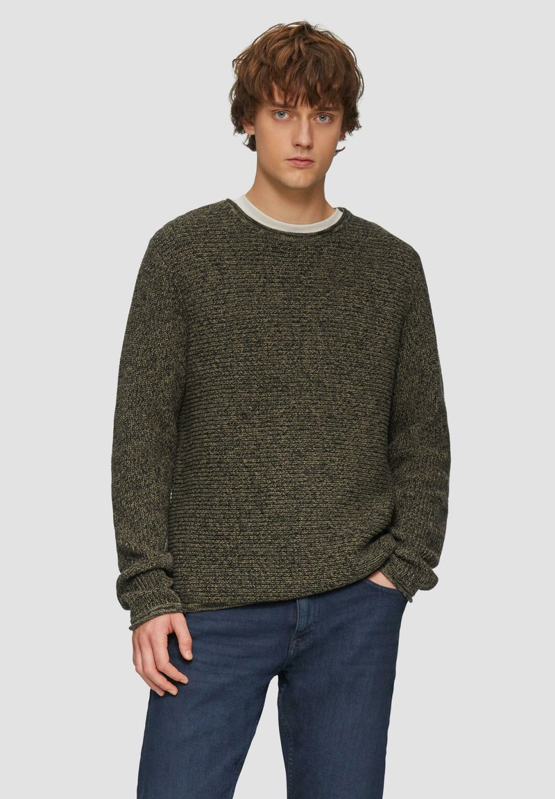 Donkergroen gestructureerd sweater met een ronde hals en lange mouwen, gecombineerd met blauwe jeans. De sweater heeft een grof gebreid patroon.