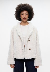 Λευκό, αφράτο, υφασμάτινο μπουφάν με γιακαδάκι και δύο καφετί κουμπιά. Χαλαρή εφαρμογή, σε συνδυασμό με ένα ανοιχτόχρωμο μπλουζάκι και σκούρα τζιν.